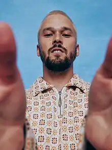 Justin Nozuka wearing a patterned shirt, standing in front of a blue backdrop, grinning and looking down at camera, which he appears to be holding in both hands