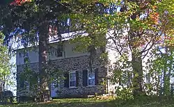 A stone house with wooden siding on the upper story, behind two large trees, one of which has some leaves turning red