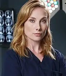 Woman with blonde hair and dark blue scrubs looking directly at the camera with a straight-faced expression