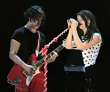 The White Stripes standing on stage: Meg is to the right, wearing a white polka dot shirt and black pants, singing into a mic; to her right is Jack, wearing a black shirt and red pants.