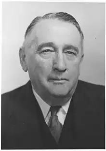 A black and white head and shoulders photograph of a balding male in a suit