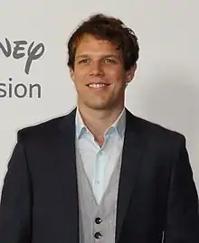 A brown-haired man wearing a suit and smiling at the camera