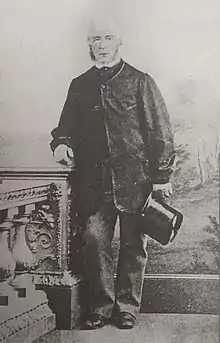 A black and white standing portrait of a white male holding a top hat