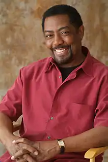 A man with dark hair wearing a white T-shirt including a silver necklace.