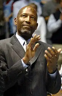 A man, wearing a gray suit, a shirt and a tie, is clapping his hands while looking up.