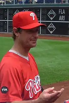 A man in a red baseball jersey with "Phillies" and red baseball cap with a white "P" on the front reaches his hand toward an unseen person.