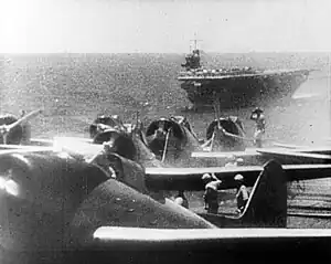 Photograph showing four aircraft lined up with several men working nearby. The sea and a complete ship are visible in the distance, along with the horizon and some sky.