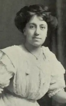 A white woman with dark hair in an updo, wearing a white dress with puffy sleeves