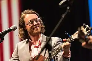 A man in a pink-and-white striped shirt and gray jacket, with neck-length hair and clear-frame glasses, stands before a microphone playing an acoustic guitar. He faces to the viewer's right. In the background on the left are the red and white stripes of a vertical American flag.