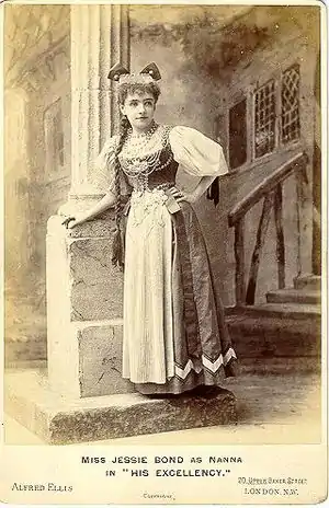 A costumed Bond, with a long braid, leaning against a column on stage.