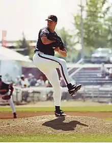 A white man in a baseball uniform in a pitching windup. His body in turned towards the camera but he is looking forward. Wearing white pants with a stripe on the left leg, his left leg is in the air. He is dressed in black shoes with a white check mark, as well as a dark jersey and baseball cap.