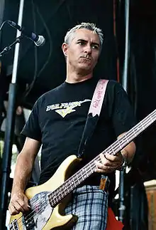 A color photograph of Joe Escalante, who is playing a bass guitar outside.