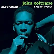 Coltrane leans back with a reed in his mouth in a deep blue-on-black photo. The words "BLUE TRAIN" are written above his head in white followed by "john coltrane" in green.