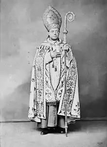 A bespectacled man holding a crosier in his left hand and wearing a mitre and liturgical vestments faces forward