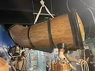 Tapered one person submarine like wooden vessel with sealed holes for operator's arms held aloft by rope slings in a museum display