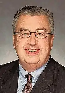 An older man with thin gray hair and glasses smiling, wearing a jacket and tie, against a gray background