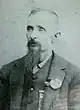 Head and shoulders of a white man with a long goatee, wearing a vest and suit coat with a flower pinned to the lapel.