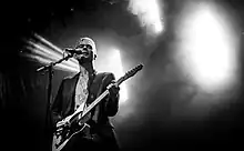 Johnny Blake stands in front of a microphone while holding a guitar