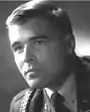 Head of a young man with neatly combed hair wearing, over a shirt and tie, a military jacket with a braided cord over his right shoulder.