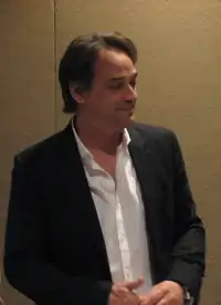 A man with dark hair, wearing a suit, including a purple tie and blue and white shirt.