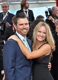 A man wearing a suite with a blue tie also accompanied with a blond hair woman wearing a black outfit.