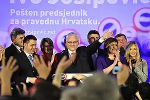 Grey-haired man with glasses, surrounded by cheering supporters