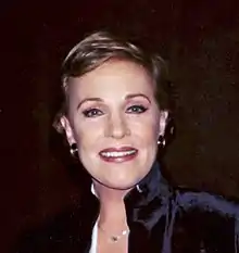 A Caucasian female with gold earrings, blue eyes, and a necklace in a navy blue outfit. She is standing against a black backdrop.