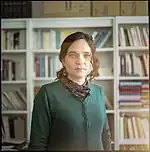 Portrait photograph of a women with brown hair and a green sweater standing in front of a bookshelf filled with books.