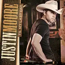 A man wearing a beige cowboy hat, black t-shirt and jeans, leaning back on a pool table.