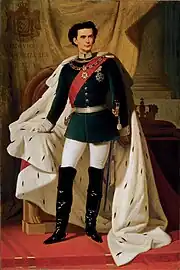 A young man in a dark military jacket, jodhpurs, long boots, and a voluminous ermine robe. He wears a sword at his side, a sash, a chain and a large star. Mainly hidden by his robe is a throne and behind that is a curtain with a crest with Ludwig's name and title in Latin. To one side a cushion holding a crown sits on a table.