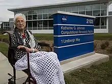  Johnson seated on a bench beside a sign in front of the Katherine G. Johnson Computational Research Facility.