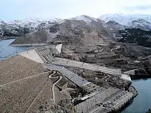 Aerial view of a concrete structure located between mountains that has a long ridge, a road and large pipes and an electric power transformation station