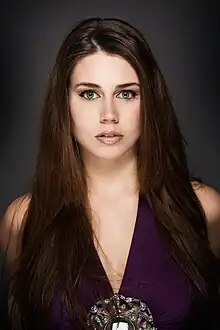 Photographic portrait of a young woman, looking directly to camera. She has long brown hair and is wearing a purple dress.