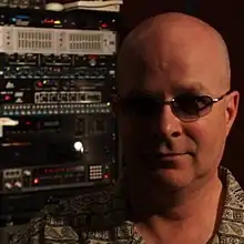 bald white male wearing shaded glasses, juxtaposed with a stack of musical equipment