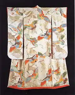 A white satin long-sleeved kimono viewed from the back. The kimono is lined in red silk with a padded skirt and sleeve hem, and is woven with a large, figured pattern of cranes in flight, and is decorated with a dyed and gold foil pattern of cranes in flight of the same size, depicted in a number of colours.