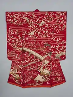 A scarlet red kimono viewed from the back, with long sleeves and a padded, lighter red hem. The kimono is decorated with cherry blossoms, traditional Japanese houses, bridges over flowing water, pine trees, reeds, clusters of bamboo leaves and small huts. The bridges, houses and water designs are depicted in couched fold thread; the other motifs are depicted in shades of pink, gold, green and white.