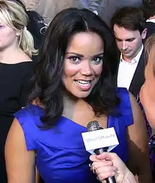 A dark-skinned woman in a blue dress talking into a microphone being held by another person