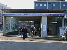 Beneath a blue sign reading "KING'S CROSS ST. PANCRAS UNDERGROUND STATION several people exit from an entrance leading to steps down. To the right there are silver doors under a sign reading "Lift". The sky is overcast, above the entrance is a dark blue temporary building