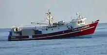 A small red fishing boat, without its usual tackle but with shipping containers at the rear, riding low astern in the open sea.