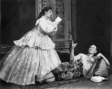 A man with a shaven head, wearing Asian dress, reclines on the floor and gestures at a woman in 19th century dress, who is writing, apparently at the man's dictation.