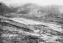 Panoramic view of a cleared strip of land surrounded by thick scrub and jungle. Behind the cleared strip the ground rises steeply