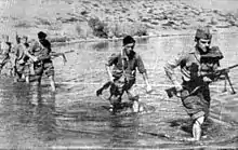 a black and white photograph of a line of males in uniform carrying weapons wading through a shallow river