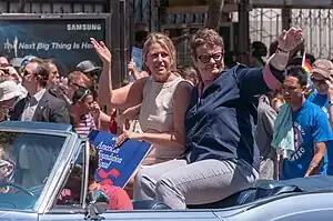 Two women sitting on the top of the back seat of an open-top automobile waving to a crowd.