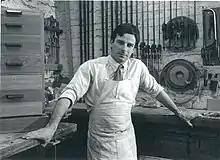 A man in a work apron stands in front of a woodworking station and facing the camera.