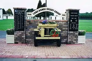 Front end of a jeep, sticking out of brick wall with plaques on brick pillars either side