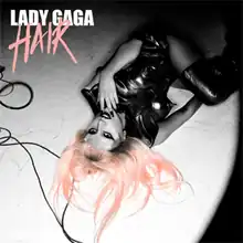 Greyscale upturned image of Lady Gaga lying on the ground, wearing a black leather dress. Her left hand is near her mouth and she looks up to the camera. On top-right the words "Lady Gaga" and "Hair" are written. Her flowing hair and the word "Hair" are colored pink.