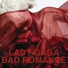 Upper bust of a woman with short cropped hair. Her body and her face is covered by a red translucent cloth with intricate wrappings in the front. Over the image the words "Lady Gaga" and "Bad Romance" are written in red capital letters. The photograph was shot by Heidi Slimane.