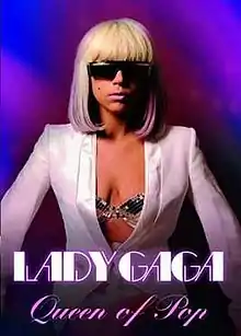 Lady Gaga in a bob cut, wearing a white coat, which reveals her sparkling bustier. She is standing in front of a blue backdrop, wearing dark shades. A black mole is visible on the woman's right cheek.