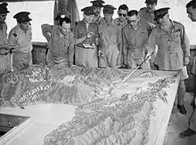 Men in military-style uniforms stand around a terrain model while one man points out terrain features with a pointer