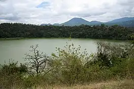 Circular volcano crater lake with sharp tree covered cliff banks and other volcanos in background.